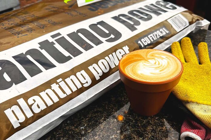 A bag of planting power soil on a counter next to a latte in a clay pot mug and gardening gloves.